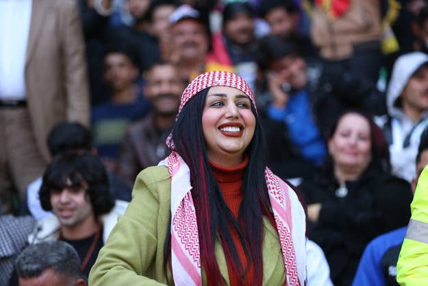 Om Fahad smiling and wearing a traditional headscarf and red sweater at an outdoor event with crowd in the background