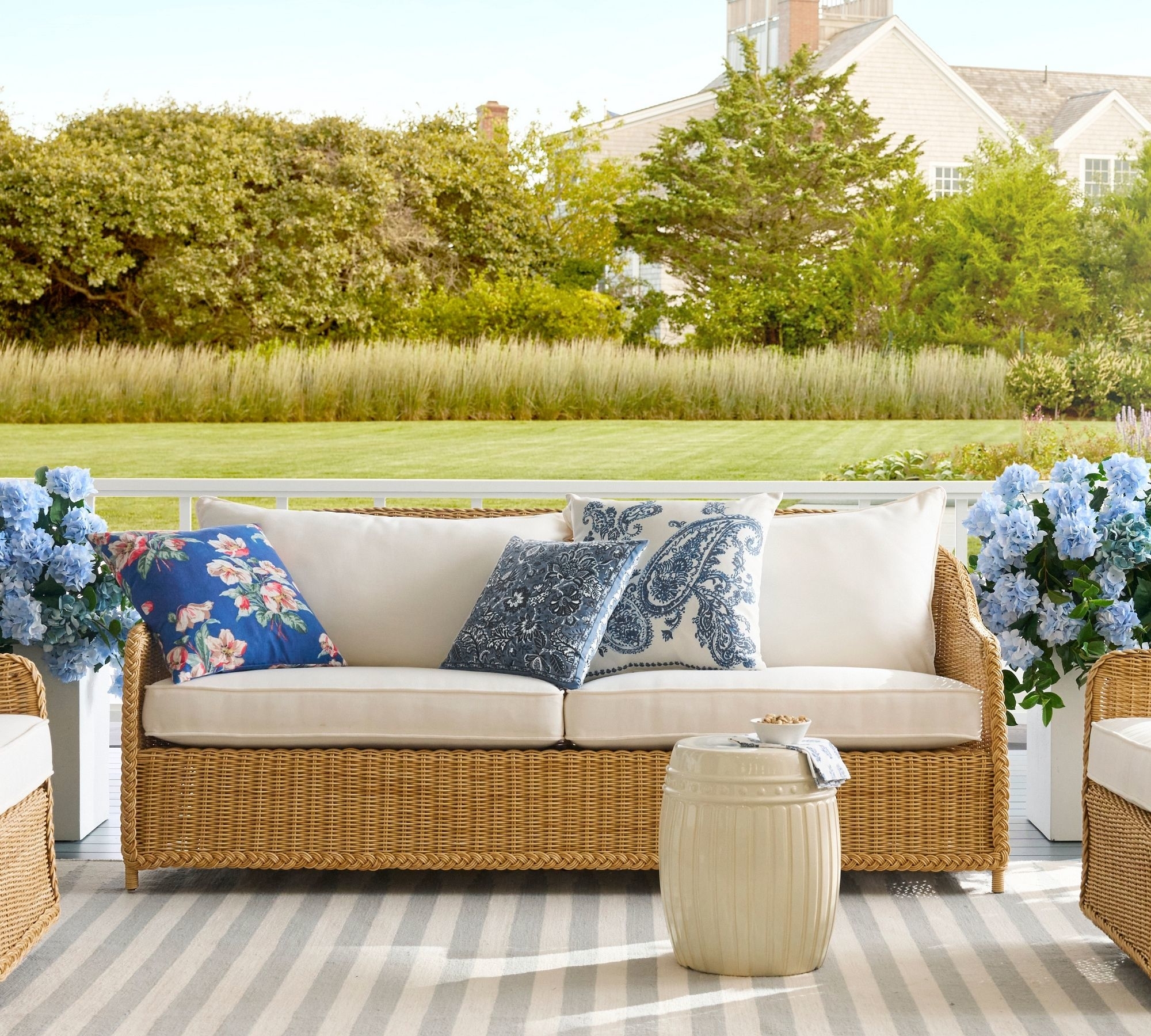 Outdoor furniture setting with a wicker sofa, decorative pillows, and a small table, on a striped rug with greenery in the background