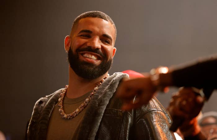 Drake smiling in a jacket and layered necklaces at a music event