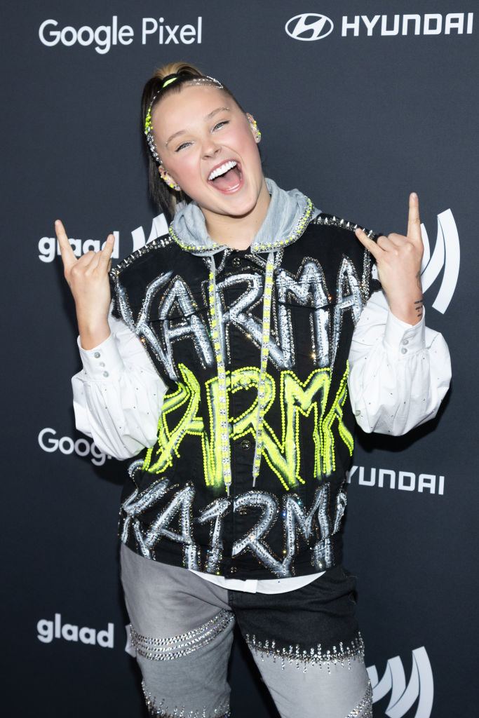 JoJo Siwa poses with a peace sign, wearing a jacket with &quot;KARMA&quot; and sparkly trousers