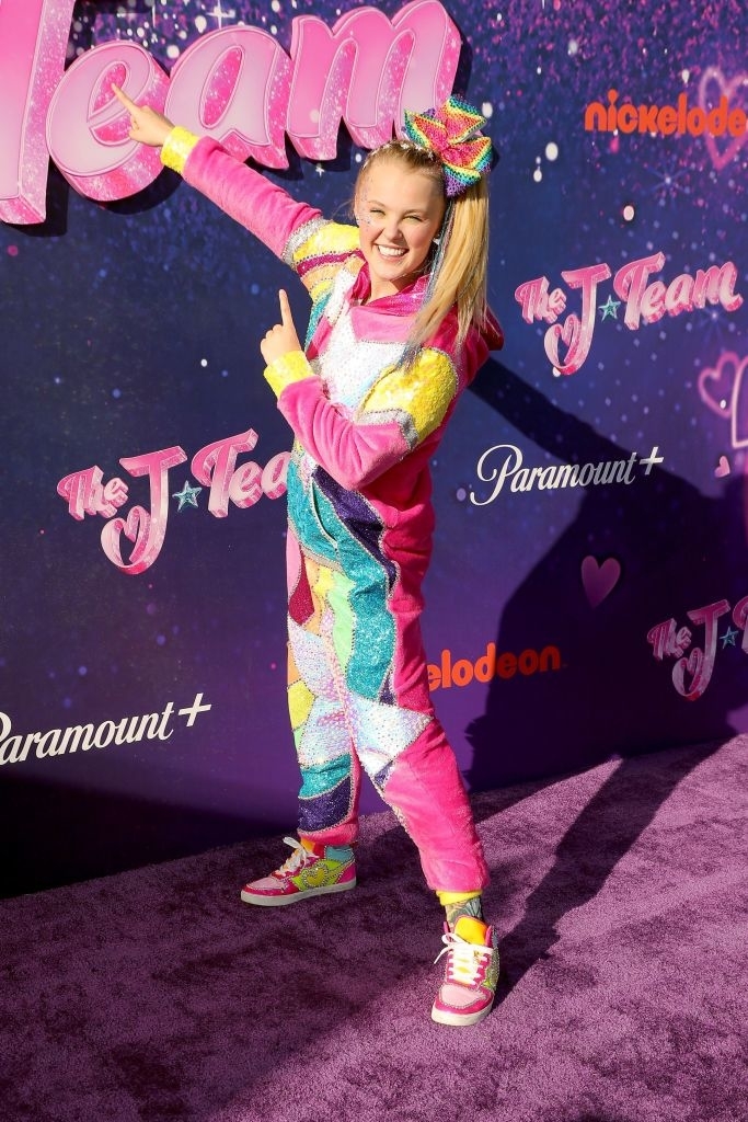 JoJo Siwa poses joyfully in a colorful outfit with a bow at a promotional event