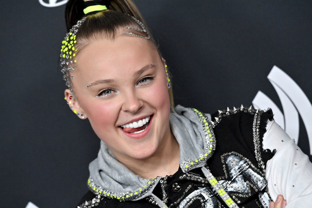 JoJo Siwa posing with a smile, wearing a sequined jacket with a hood and hair styled with vibrant hairpieces