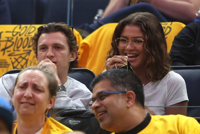 Tom Holland and Zendaya sitting together, smiling at a basketball game, among a crowd of fans