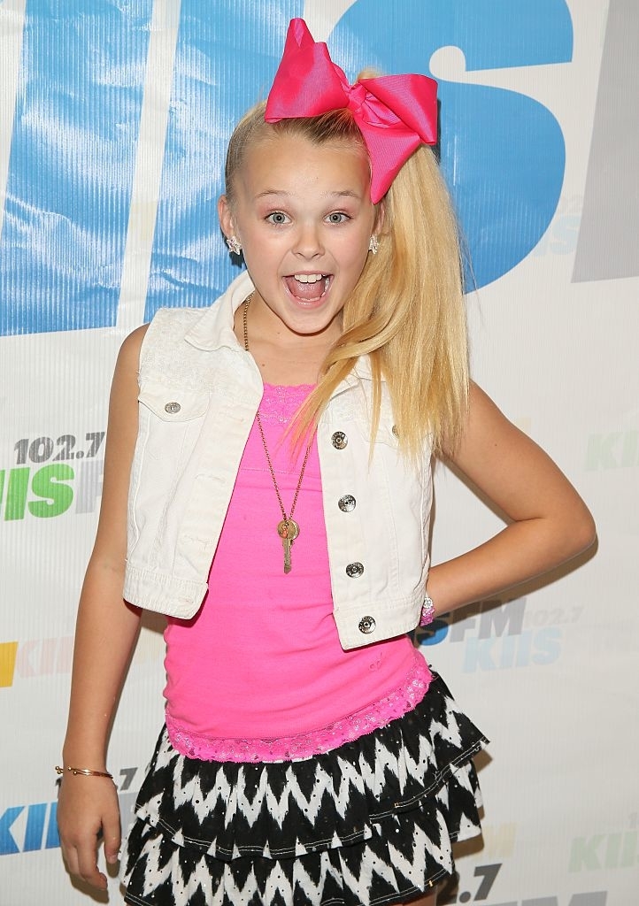 JoJo Siwa poses with hands on hips, wearing a large bow, vest, and layered skirt