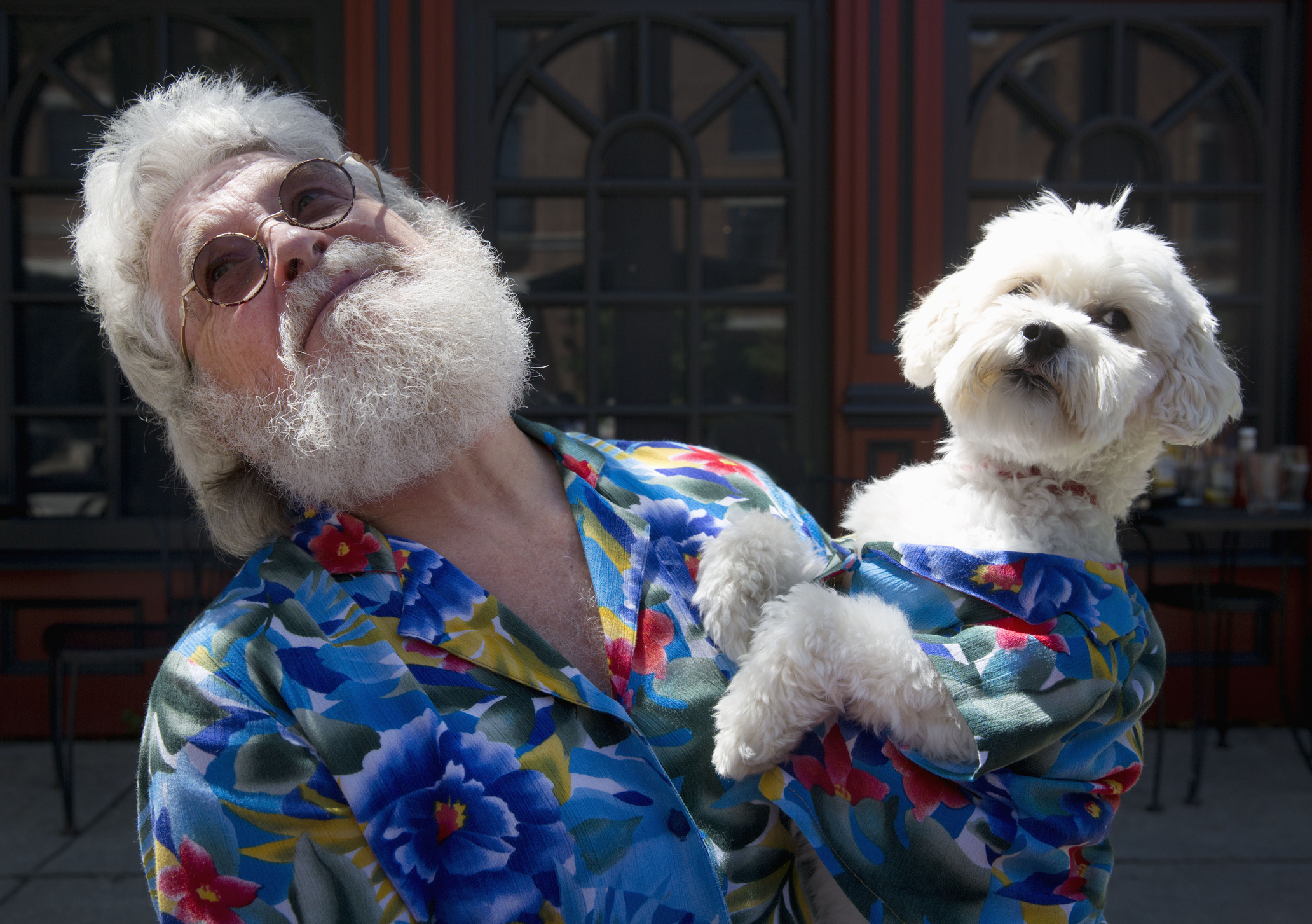 Man in floral shirt holding a small dog, both gazing upwards. They are outdoors near a building