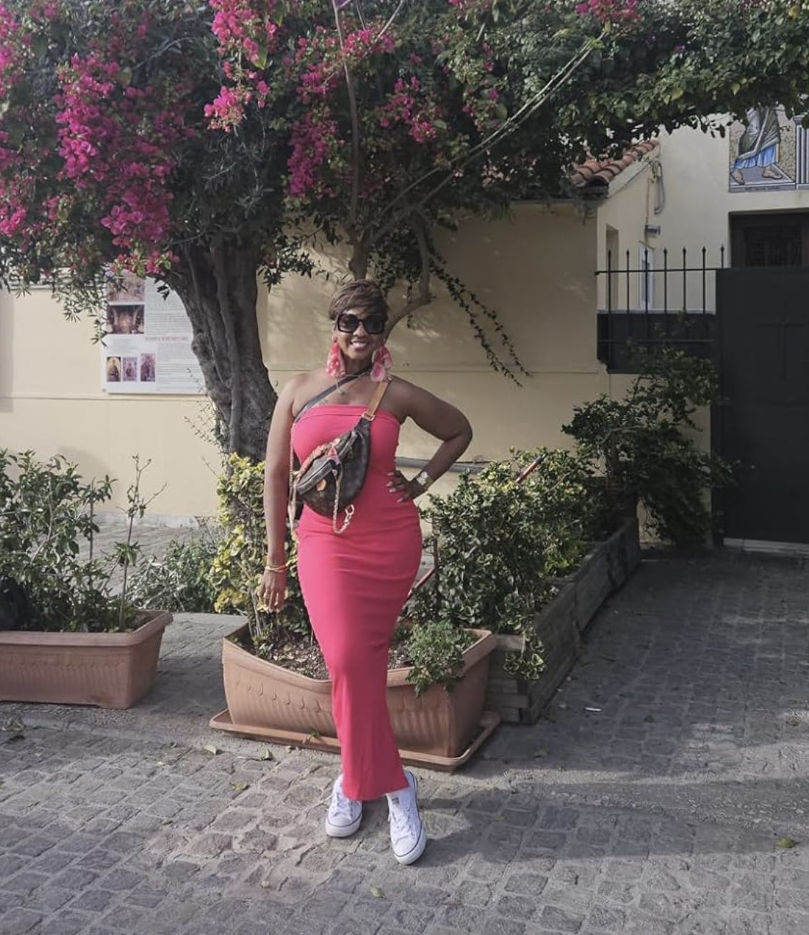 reviewer successful  a pinkish  sleeveless formal  poses with manus  connected  hip, accessorized with a loop  and sunglasses, lasting  by flowering plants
