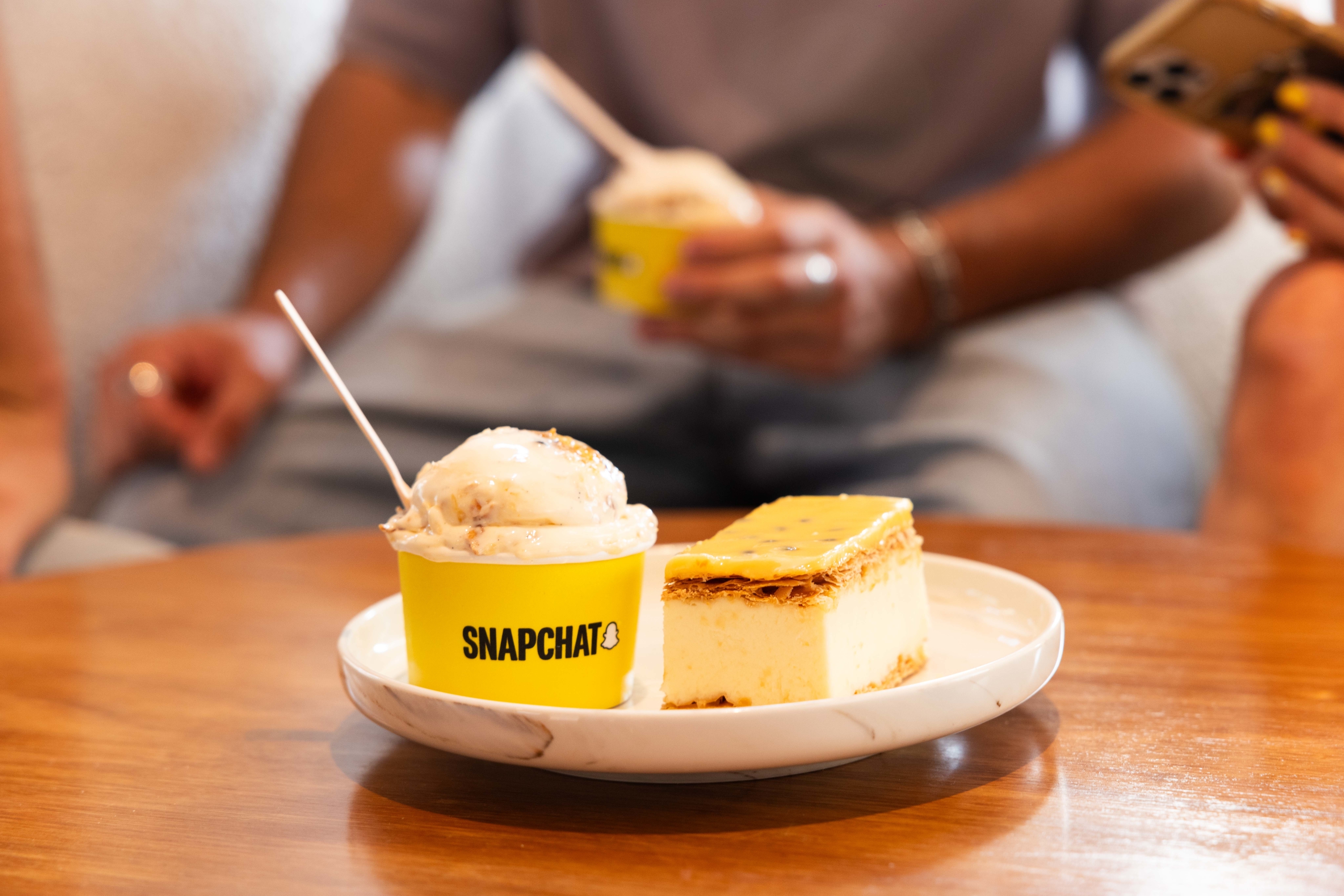 A Snapchat-branded dessert cup next to cheesecake, with people holding phones in the background