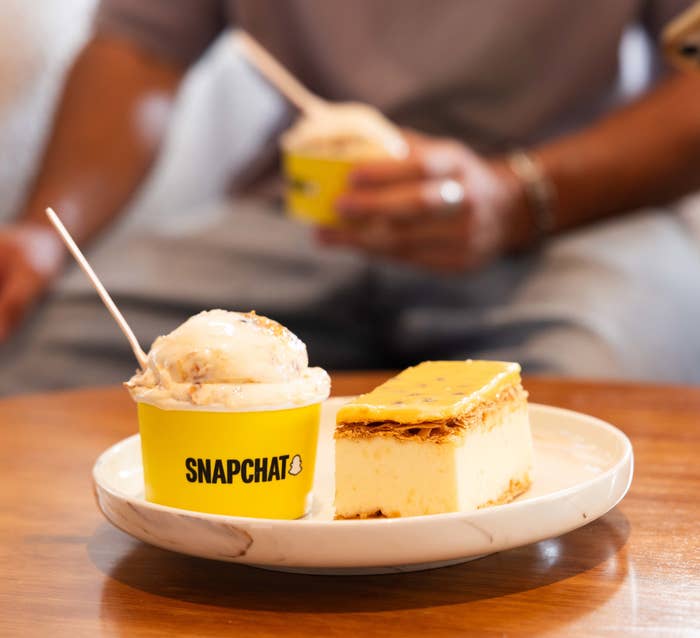 A Snapchat-branded dessert cup next to cheesecake, with people holding phones in the background