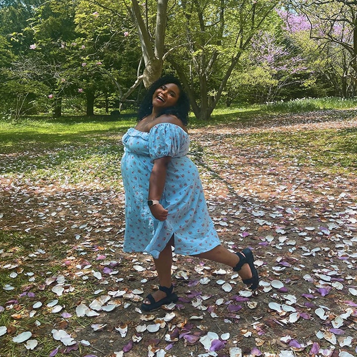 reviewer in a floral dress stands in a park with fallen petals, smiling with one hand on hip
