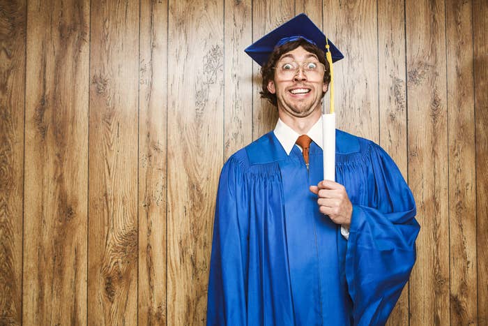 Person in graduation cap and gown smiling, holding a diploma, against a wooden wall