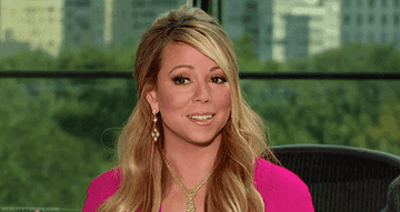Mariah Carey smiles in an interview, wearing gold jewelry and a pink top