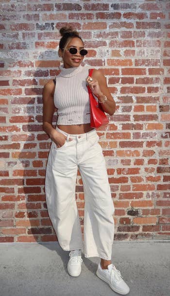 Woman in a sleeveless top, wide-leg pants, and sneakers, holding a red bag, posing in front of a brick wall