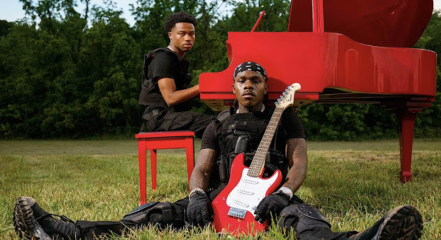 Two musicians with a red piano and guitar outdoors, one sitting on a stool, the other lying on the grass