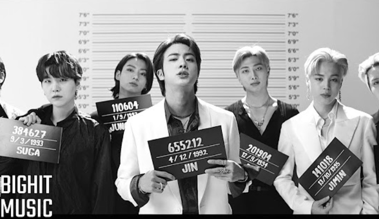 Seven members of BTS in a lineup holding signs with their stage names: V, Suga, Jin, RM, Jimin, J-Hope, Jungkook