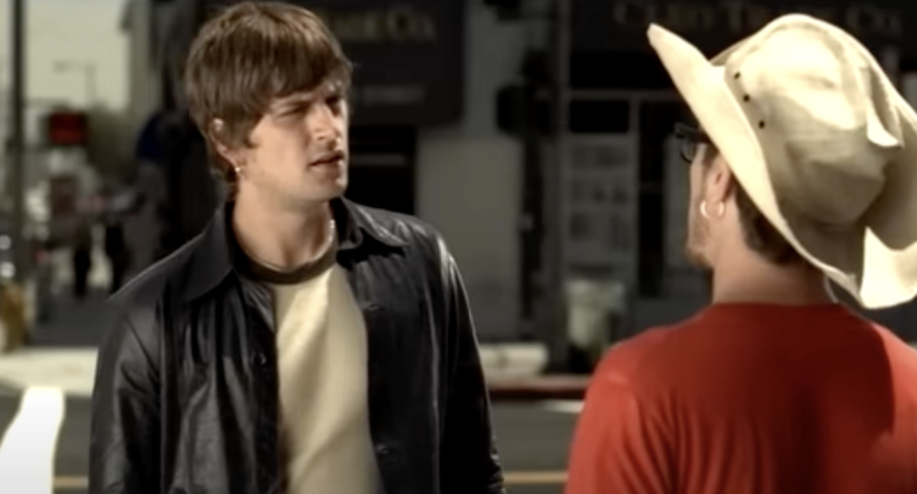 Two men talking on a street, one wearing a cowboy hat and the other in a casual jacket