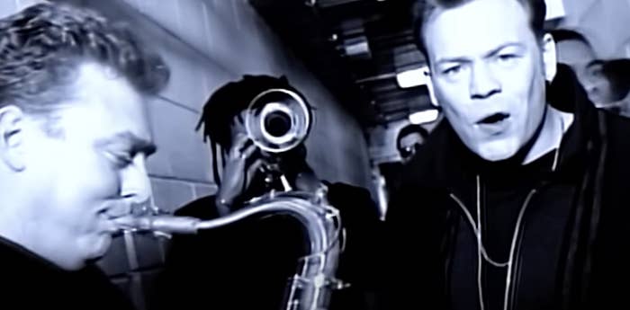Two men in a dimly lit corridor, one playing a saxophone, the other singing into a microphone