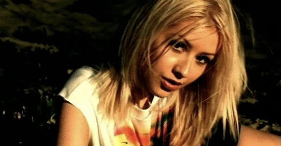 A person with blonde hair wearing a graphic tee, slightly leaning forward with a blurred background