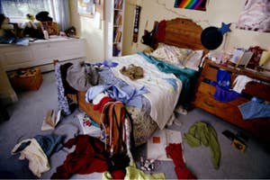 A cluttered bedroom with clothes and objects strewn about, unmade bed in center, no people visible