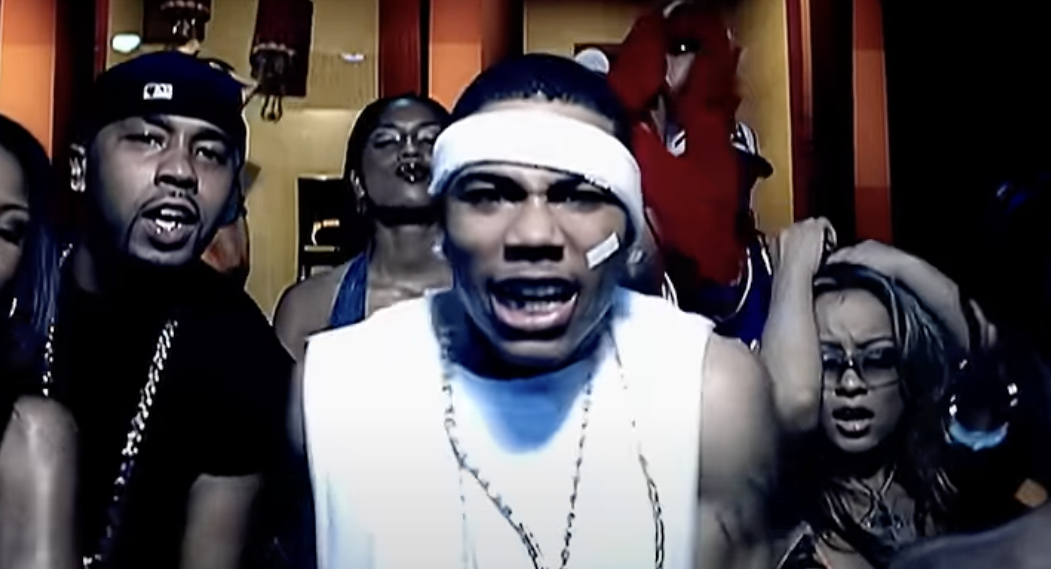 Group of people in a music video, featuring a man in a sleeveless shirt and headband centered