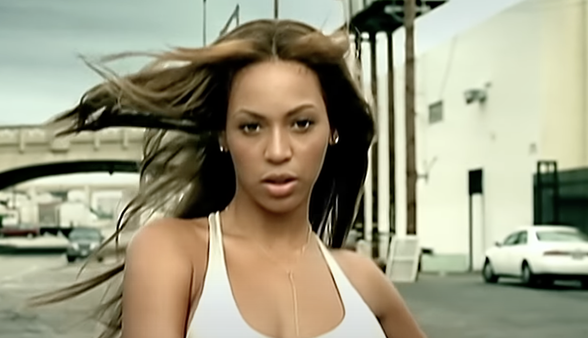 Beyoncé in a white tank top with windswept hair, stern expression, urban backdrop