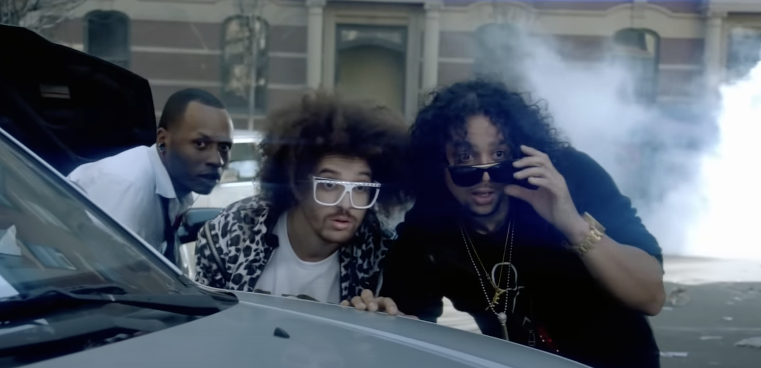 Three musicians pose by a car, one wearing a patterned jacket and sunglasses
