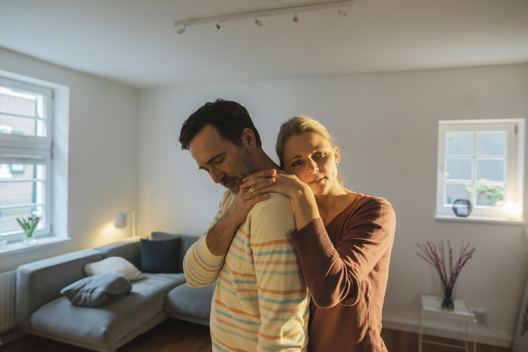 Woman embracing man from behind, both look thoughtful, in a living room setting