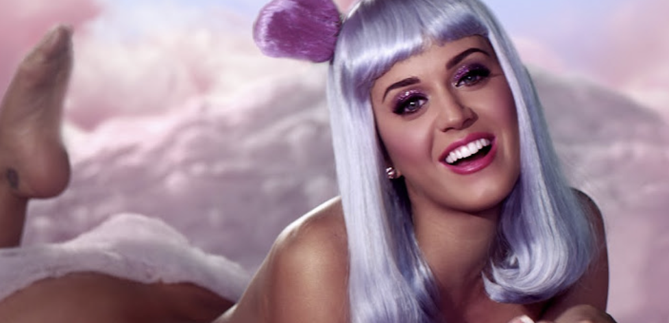 Katy Perry lying on clouds wearing a wig and whimsical makeup, facing the camera with a playful expression