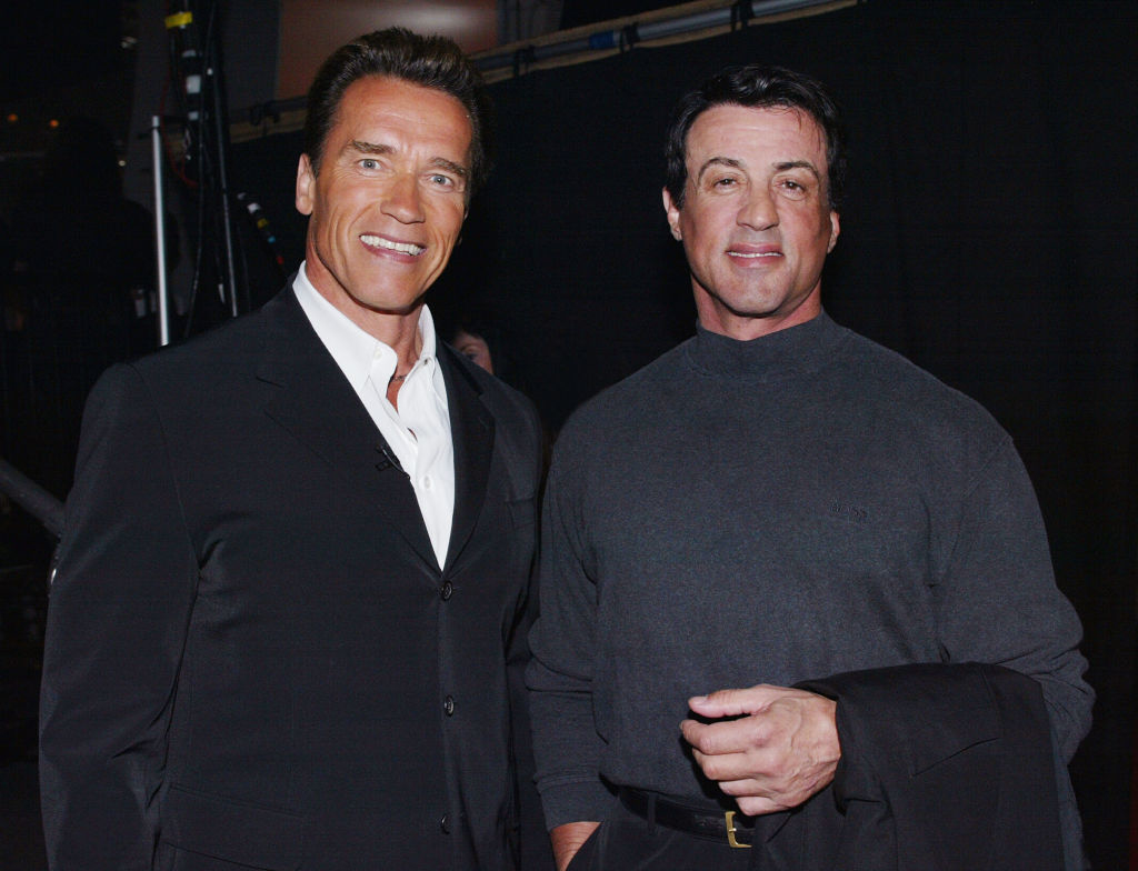 Arnold Schwarzenegger in a suit and Sylvester Stallone in a turtleneck smiling side by side