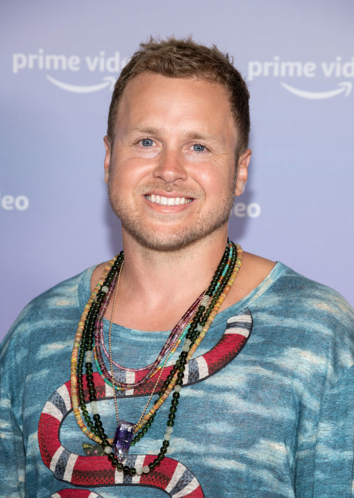 Spencer Pratt posing with a smile, wearing a patterned blue shirt and multiple beaded necklaces