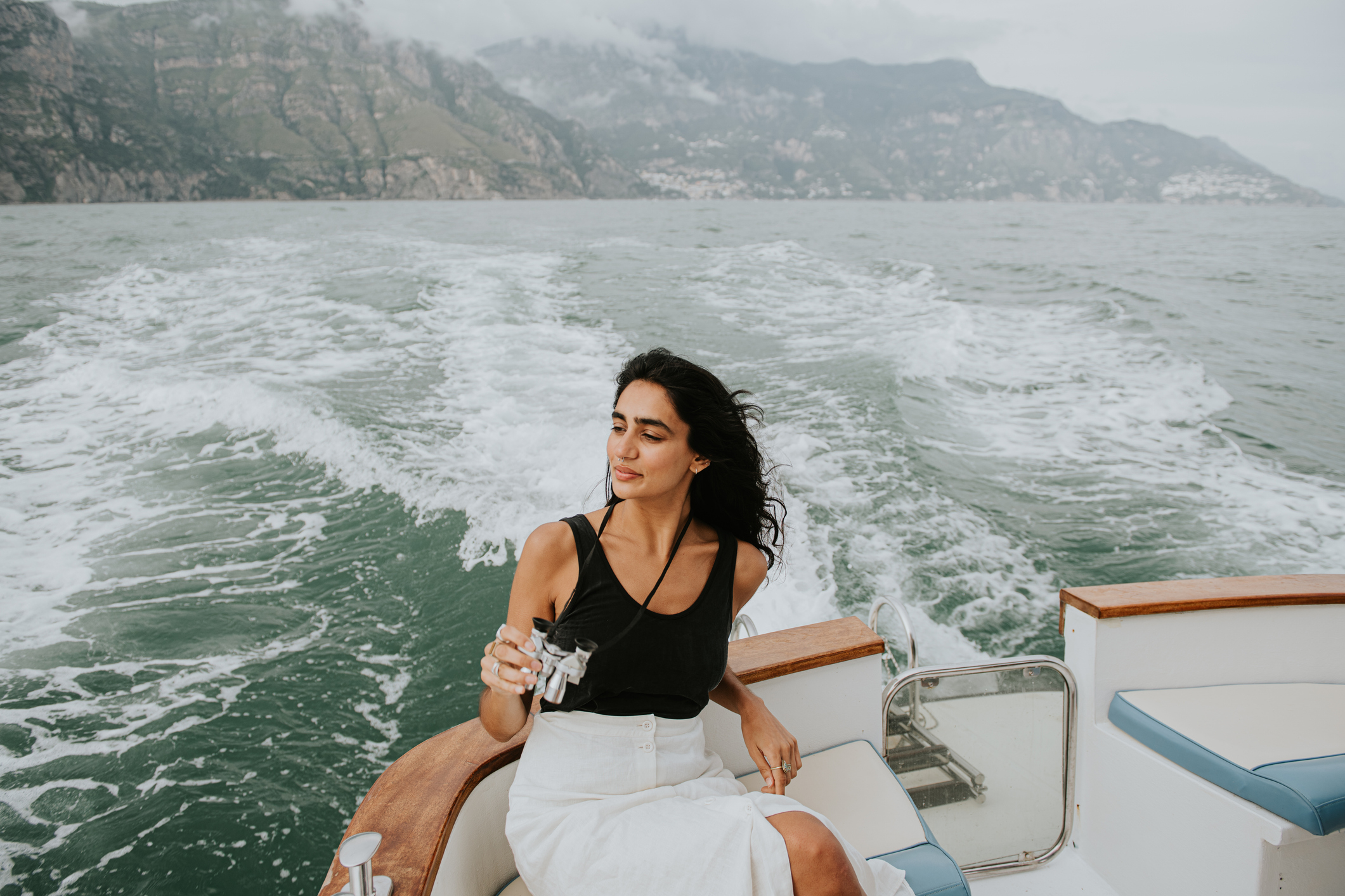 Woman relaxing on a boat with a drink, hair blowing in the wind, facing the sea