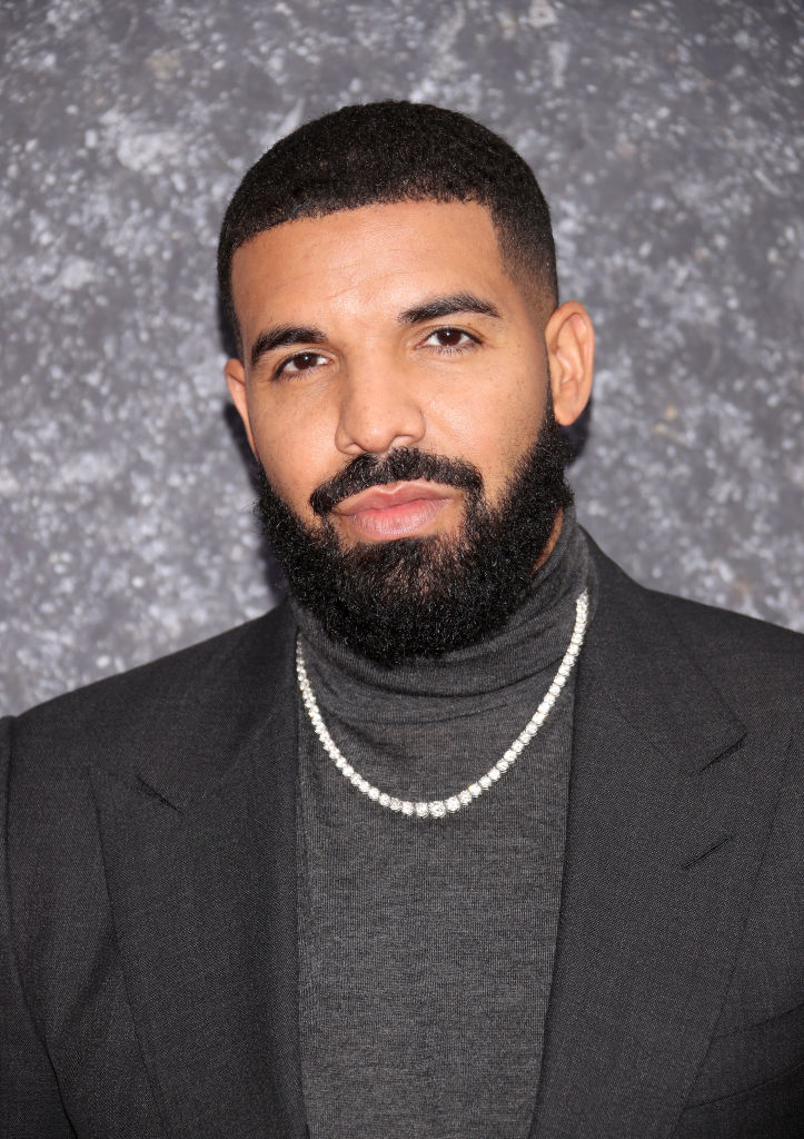 Drake in a black turtleneck and jacket, standing before a speckled backdrop with a silver necklace