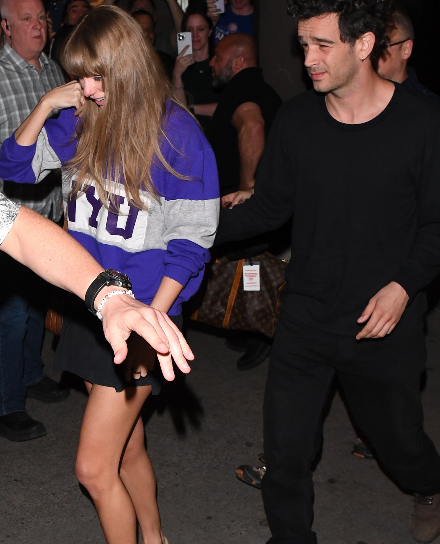 Taylor Swift in a purple sweatshirt with the number &quot;13&quot; escorted by Matty Healy in black through a crowd