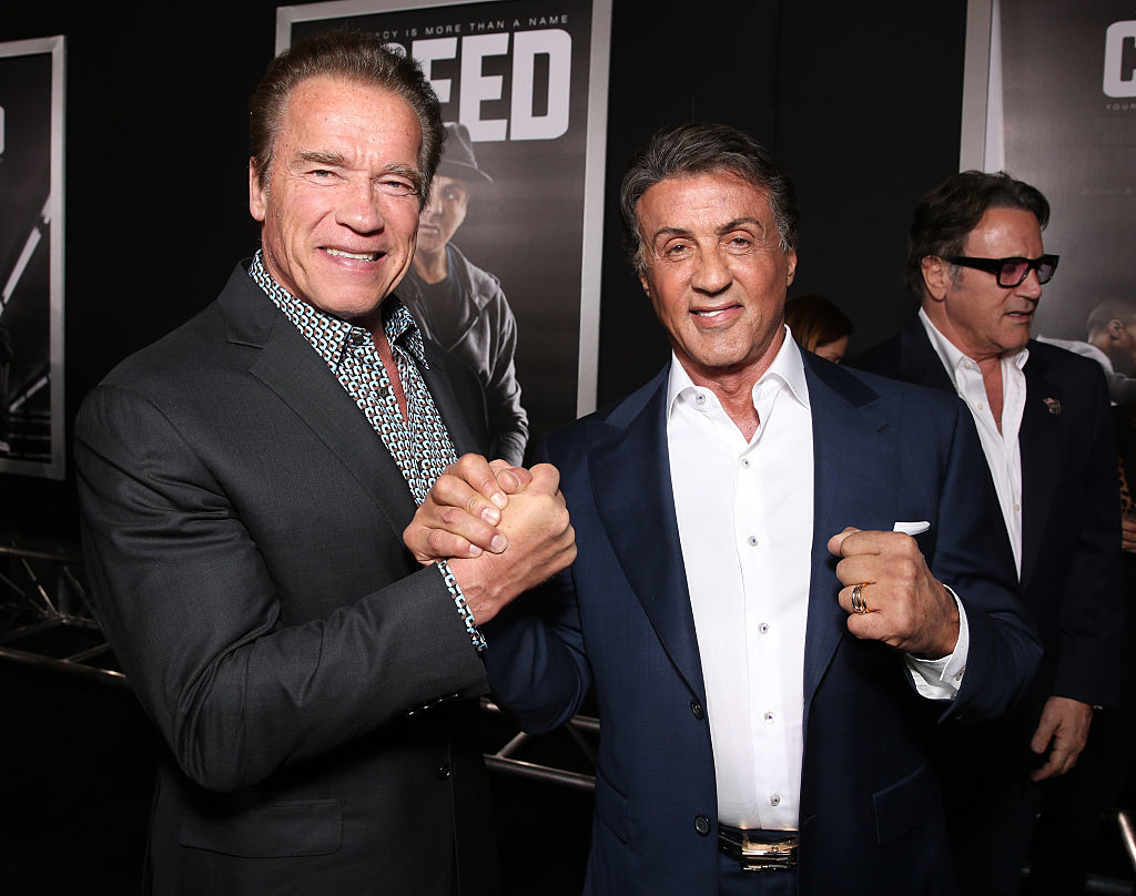 Arnold Schwarzenegger and Sylvester Stallone smiling, clasping hands at an event, both in formal attire