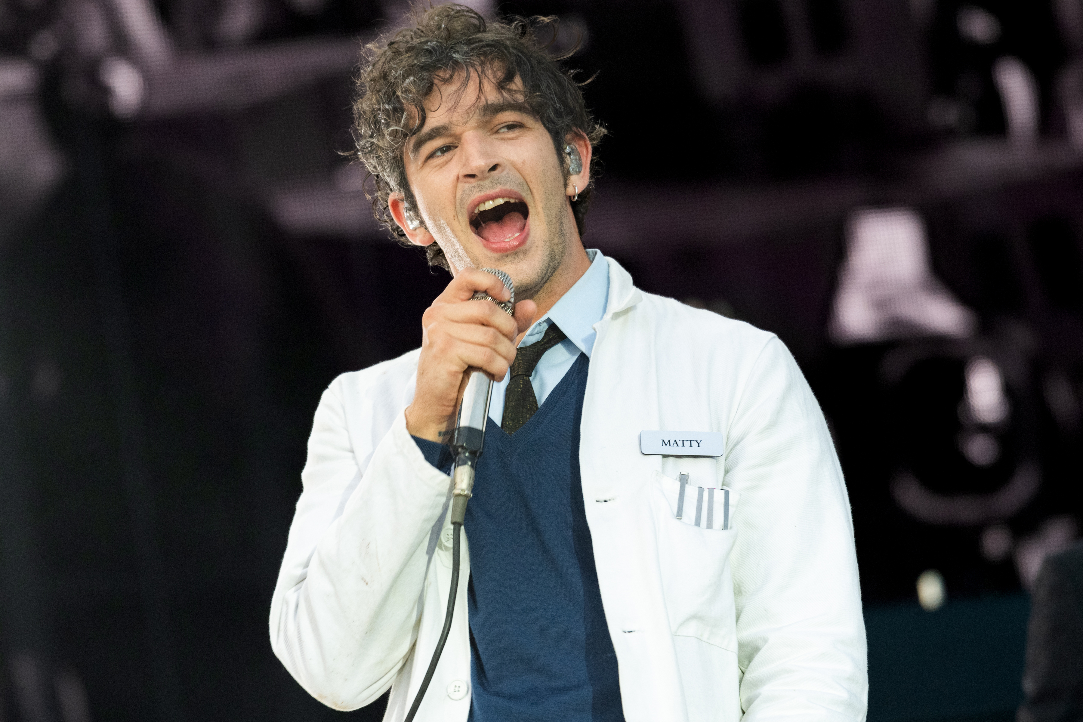 Matty Healy of The 1975 performing on stage, wearing a white lab coat with &quot;Matty&quot; name tag
