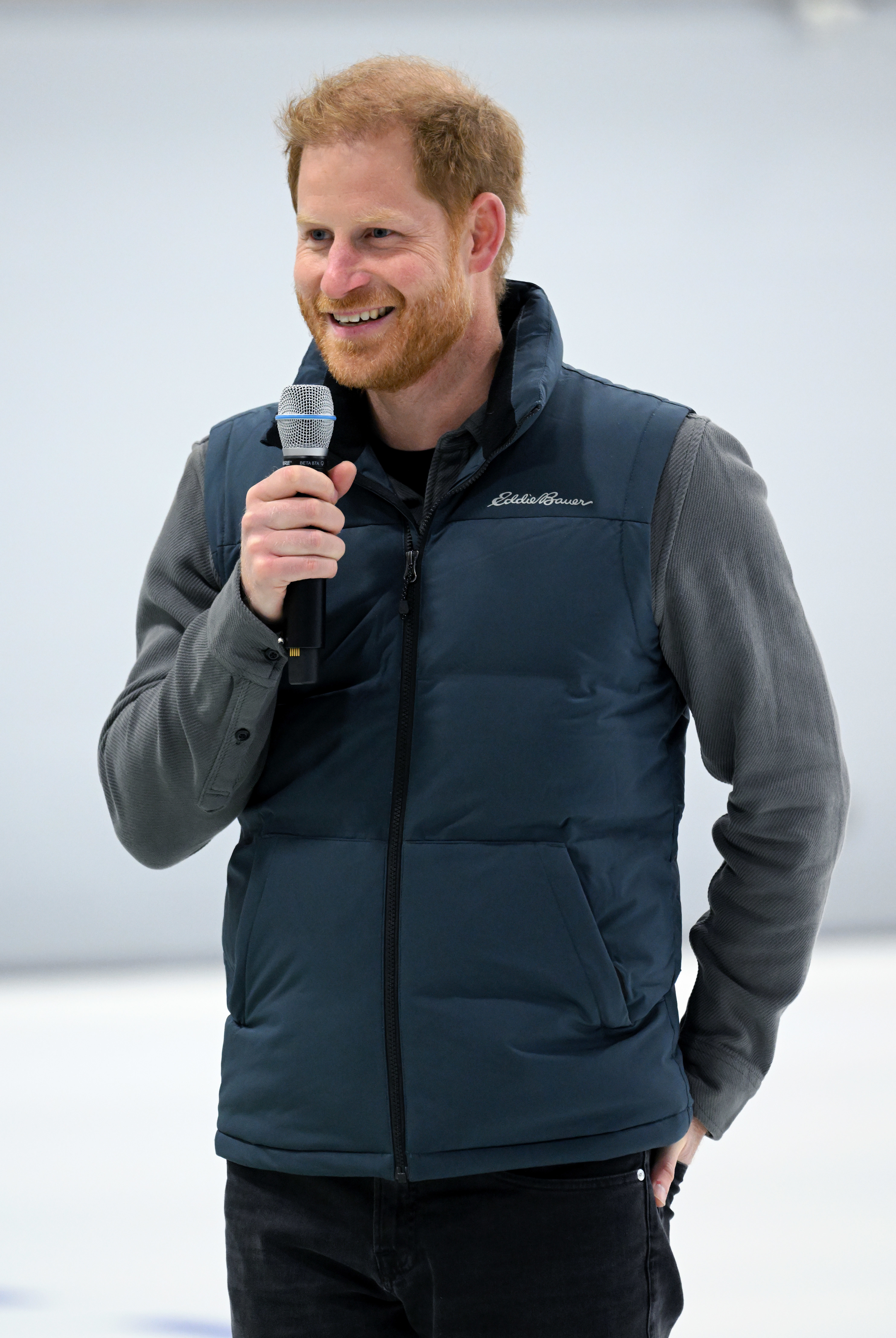 Prince Harry smiles holding a microphone, wearing a casual zip vest and long-sleeve shirt