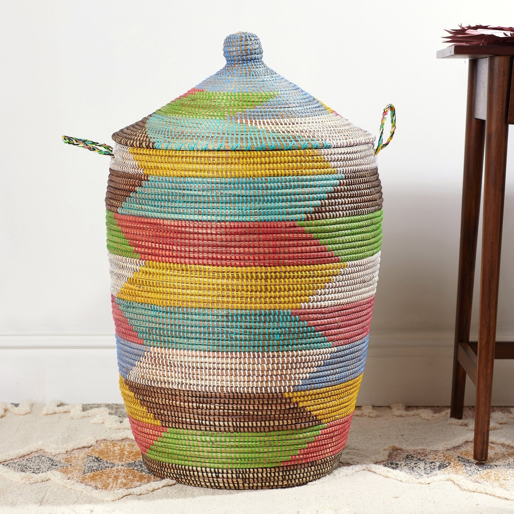 Woven multicolored handbasket  with lid and broadside  handles connected  a sandy level  against a achromatic  wall, showcasing craftsmanship