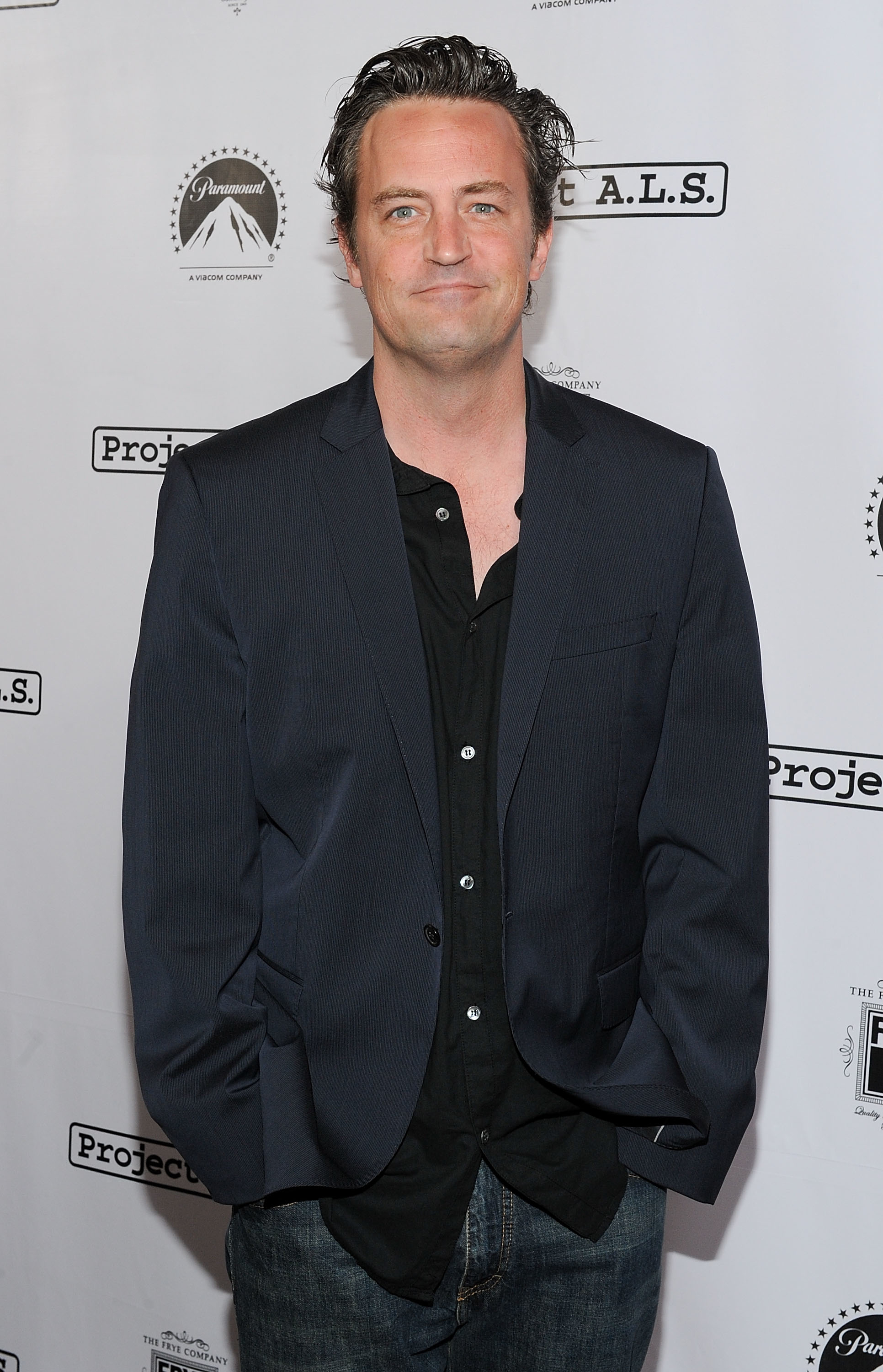 Matthew Perry stands on the red carpet wearing a dark suit with an unbuttoned shirt
