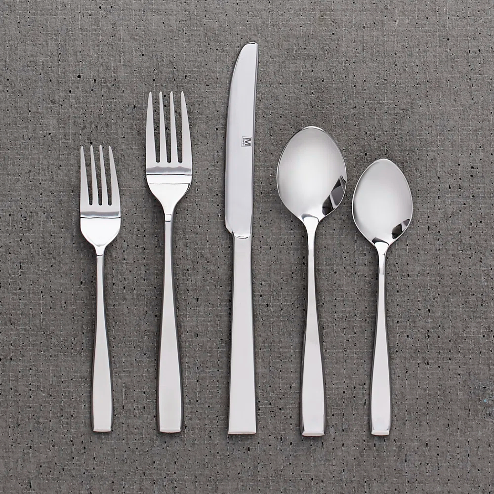 Five pieces of flatware including two forks, a knife, and two spoons are arranged side by side on a textured background