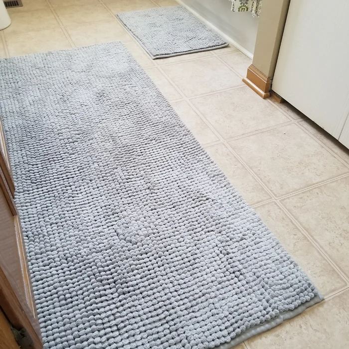 Reviewer&#x27;s photo of two of the bath mats in different sizes, both light blue, on the floor in a bathroom, one in front of the sink and the other in front of the toilet