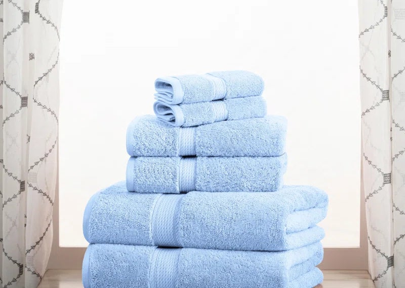 The towel set in the color blue, folded and stacked on a wooden surface with a curtained window in the background