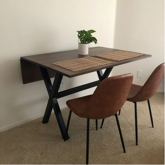 Reviewer&#x27;s photo of the dining table against a wall with one leaf extended and the other folded down, decorated with bamboo placemats, a plant, and two orange dining chairs
