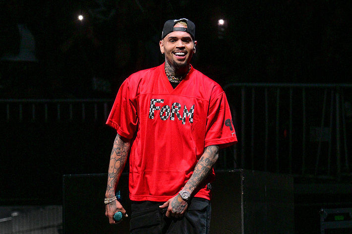 Chris Brown wearing an oversized red shirt and jewelry, holding a microphone onstage