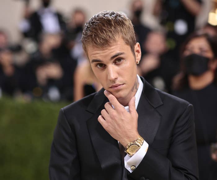 Justin Bieber in a black suit and gold watch posing with a hand on chin