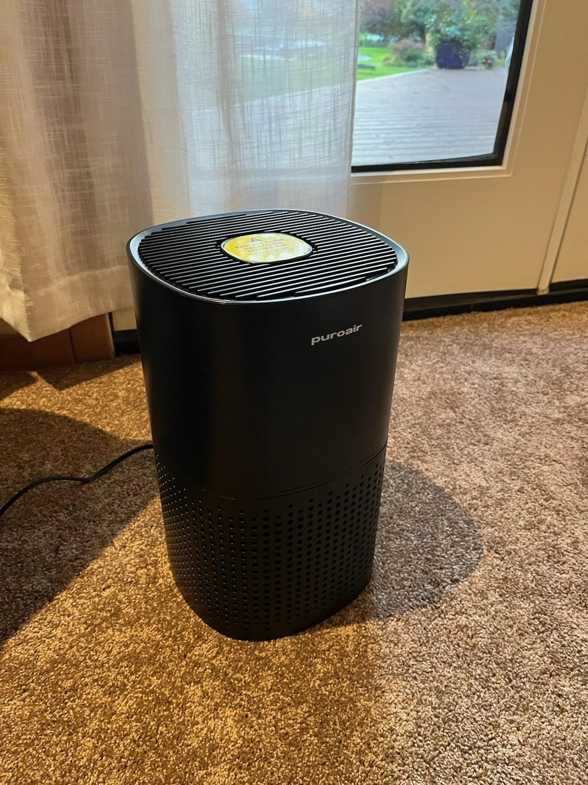 Black air purifier standing on carpet near a window with sheer curtains