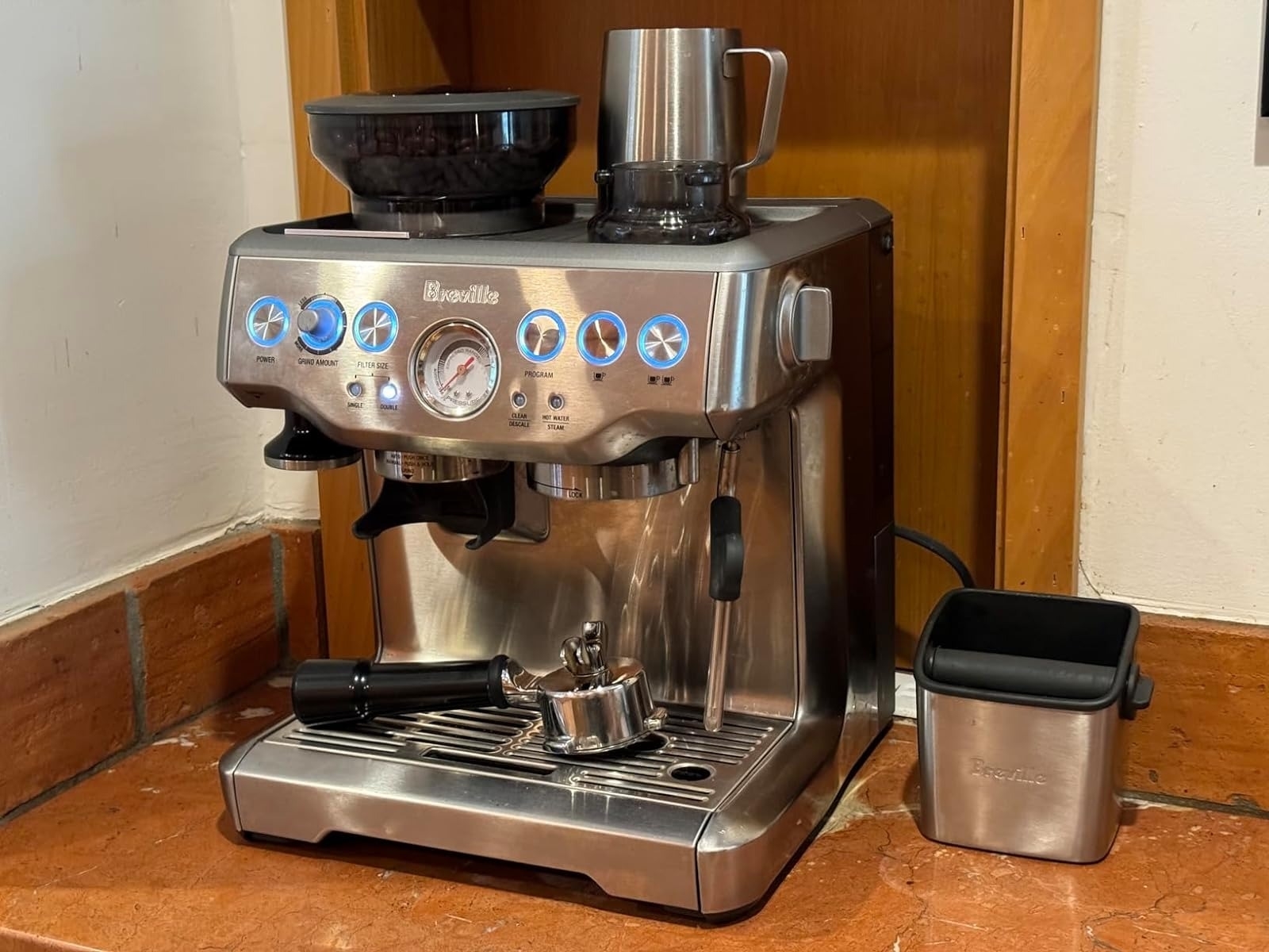 Stainless steel espresso machine with buttons and a gauge, next to a silver pitcher and black container