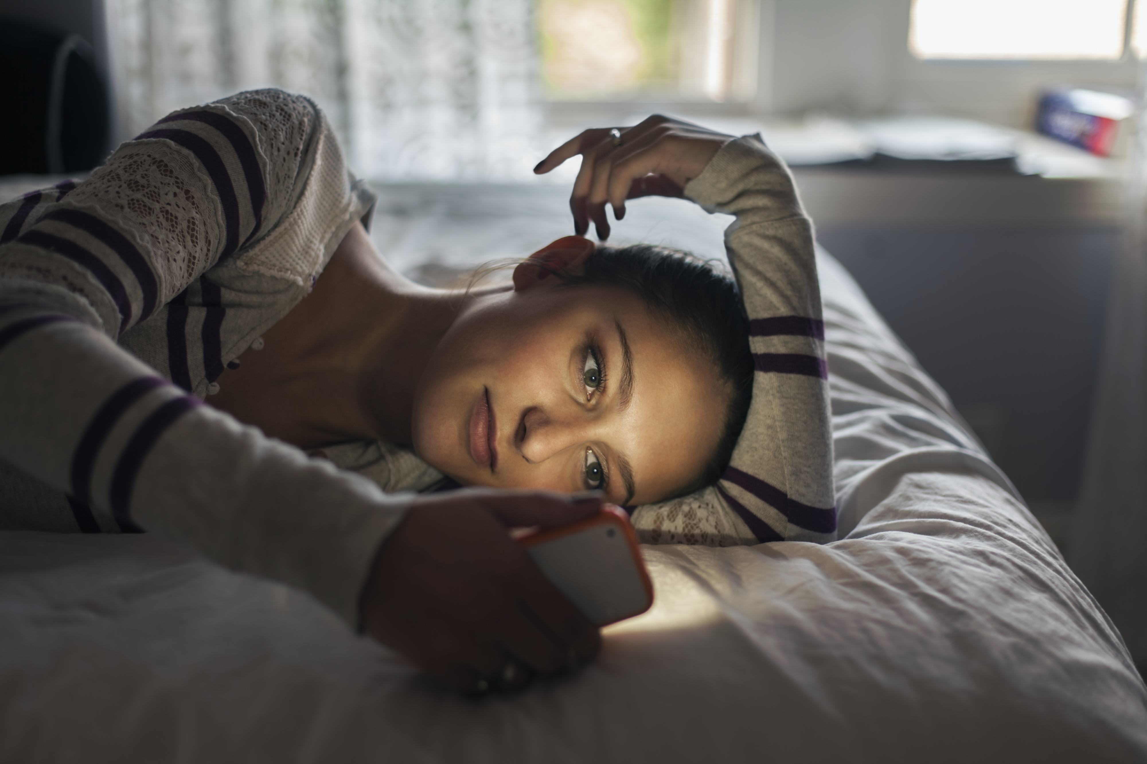 Person lying on stomach on a bed, holding a smartphone, looking pensively towards the camera