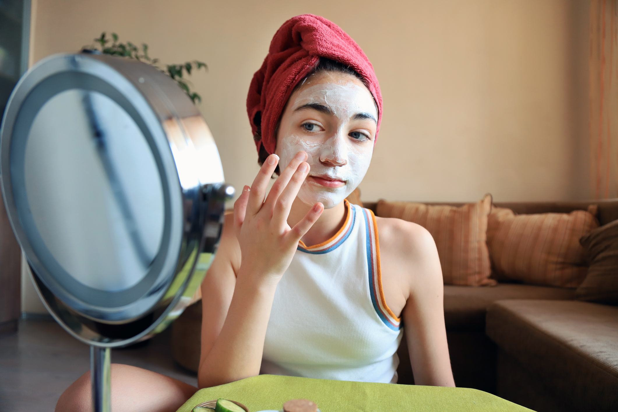 Young person applying a facial mask while looking into a mirror, dressed in casual sleeveless top with towel-wrapped hair