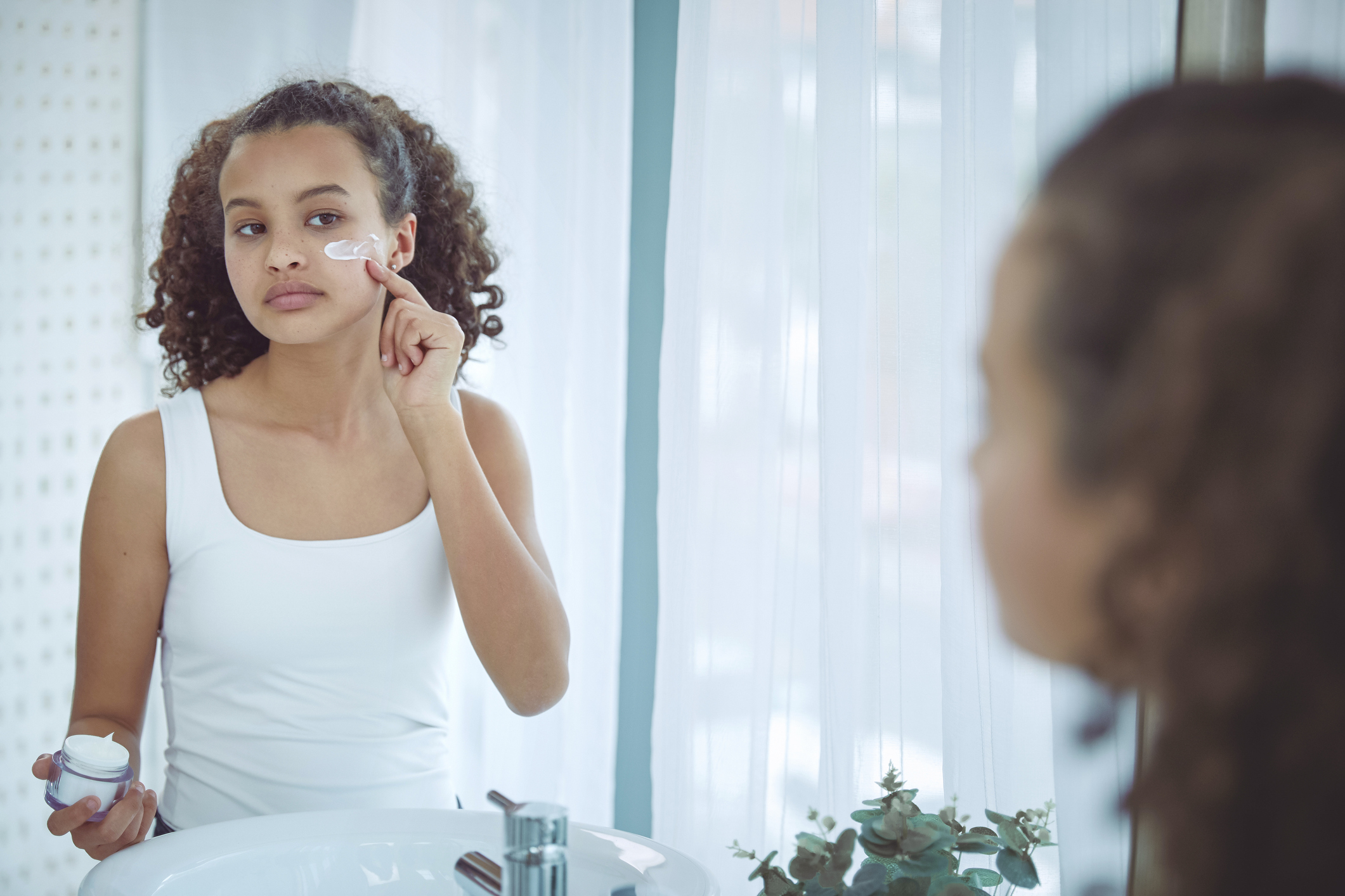 Young person applying facial cream, reflected in a bathroom mirror