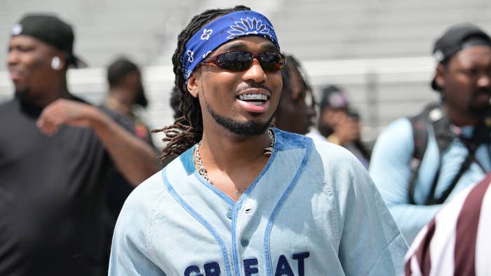 Person smiling, wearing a bandana, sunglasses, and a jersey with the word &quot;GREAT&quot; across it, amidst a crowd