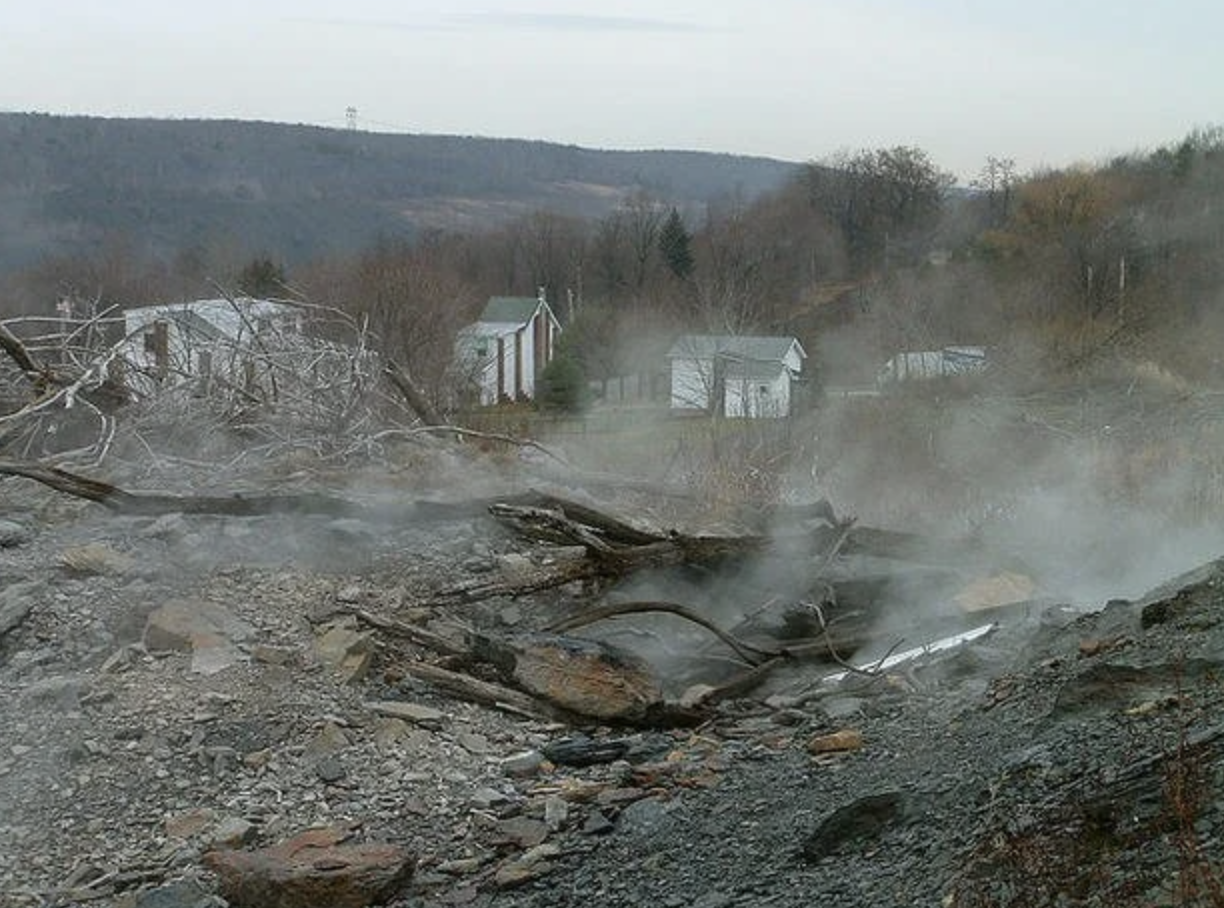 Smoke rises from the ground in a barren landscape with damaged trees and abandoned buildings in the background